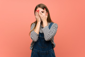 Young woman with overalls over pink wall covering eyes by hands and looking through the fingers
