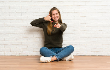 Young woman sitting on the floor making phone gesture and pointing front