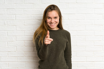 Young woman over white brick wall points finger at you