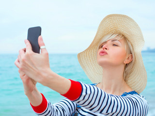 beautiful young blonde caucasian woman on vacation in straw hat, striped blouse nad denim overalls making selfie on smartphone by the amazing blue sea background