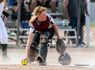 Skilled softbaall catcher with red hair and protective gear gaining a grip on the loose ball while...