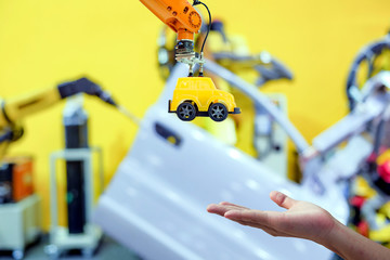 Close-up industrial robotic gripping yellow toy car send to hand human on blurred smart car factory background, industry 4.0 and iot technology concept