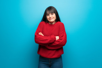 Woman with red sweater over blue wall making doubts gesture while lifting the shoulders