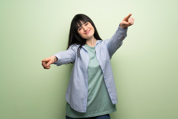 Young woman over green wall points finger at you while smiling