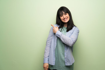 Young woman over green wall pointing to the side to present a product