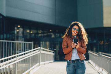 Young modern woman in brown leather jacket using smartphone in the city