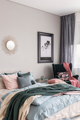 Stylish poster in black frame on grey wall of fashionable bedroom interior with emerald green armchair and king size bed