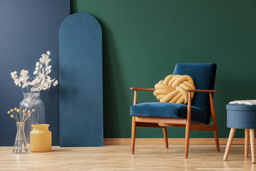 Yellow pillow on wooden armchair in blue and green flat interior with flowers and stool. Real photo