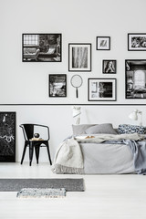 Rugs and black chair next to bed in white bedroom interior with gallery of posters. Real photo