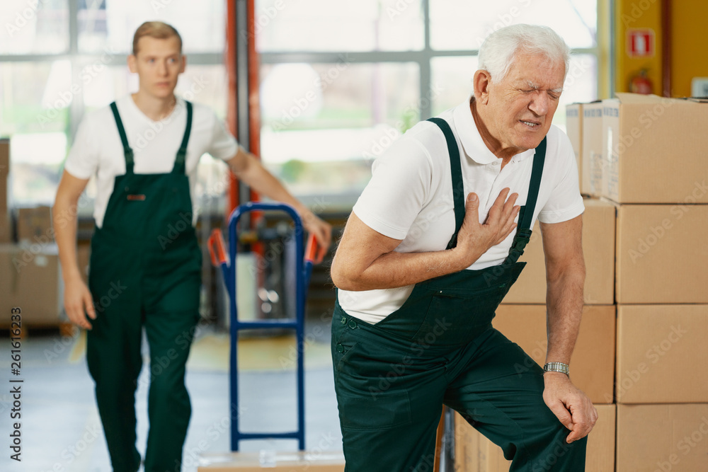 Wall mural senior warehouseman having a heart attack at work, young colleague running to help him