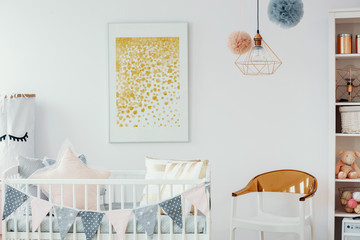 Yellow poster above cradle next to chair in white baby's bedroom interior with lamp. Real photo