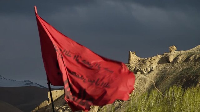 A hand held, medium shot of a big Muslim, red flag situated in the middle of the nature, moving in the wind beneath the cloudy sky.