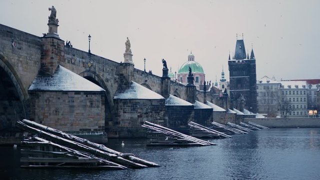 Snowy Charles Bridge in Prague, cold murky morning with lots of birds flying IN THE DISTANCE, Vltava river, kampa first snow cloudy, Czech Republic. Flock flies across the frame in front of camera at 