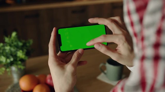 Over shoulder view of male hand actively gesturing, using horizontal green screen touchscreen of the smartphone on blurred wooden kitchen table background. Texting messages, surfing social networks