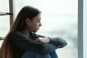 Upset teenage girl sitting at window indoors. Space for text