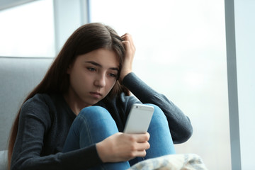 Upset teenage girl with smartphone sitting at window indoors. Space for text