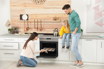Woman and her family taking out tray with baked cookies from oven in kitchen