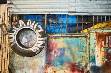 Colorful painted rusty entrance metal gate with an attached old tire as signage. Located behind the gate is an operating vulcanizing business
