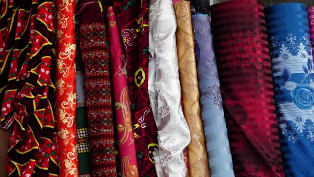 A steady, close up shot of piles of colourful fabric in a market and people passing by.
