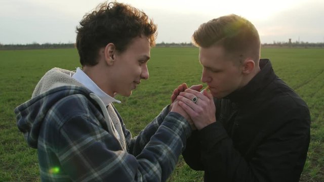 funny boy helping his friend to light a cigarette in a windy day in the green field at sunset
