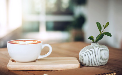 Closeup of latte art coffee on wooden table with book and small tree in ceramic vase background, vintage color tone