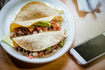Three delicious tacos on a plate sitting next to a cell phone at a Mexican food restaurant. Concept of ordering your food and lunch online