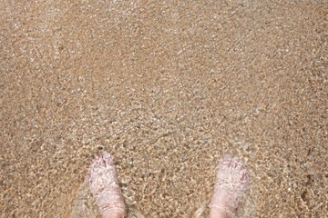 A pair of white human feet stand barefoot in shallow water as light glistens on the surface of the water.