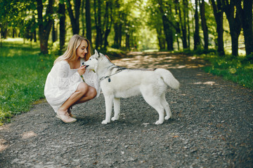 Beautiful and gentle girl with light hair dressed in white dress is playing along with her sweet doggy in summer green park.
