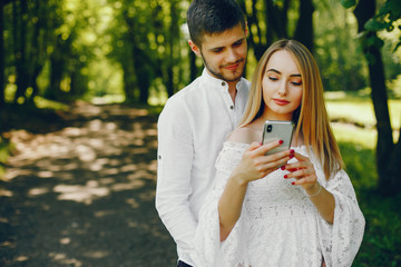 A beautiful and gentle girl with light hair and a white dress is walking in a sunny summer forest with her handsome guy in a white shirt and dark pants and they use the phone