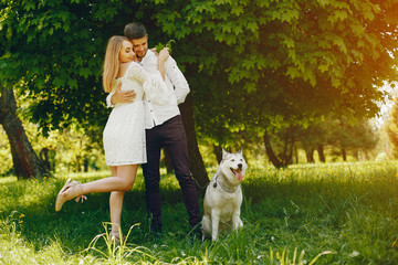 Naklejka na ściany i meble A beautiful and gentle girl with light hair and a white dress is walking in a sunny summer forest with her handsome guy in a white shirt and dark pants and they playing with cute gray dog