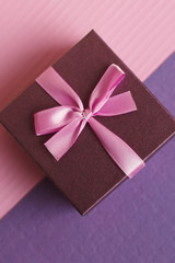 Small gift box with pink ribbon and bow on pink and purple background, vertical photo, top view