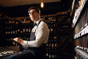 Professional male Sommelier dressed in bowtie and apron writes at notepad, putting down information on wine bottles in wine house