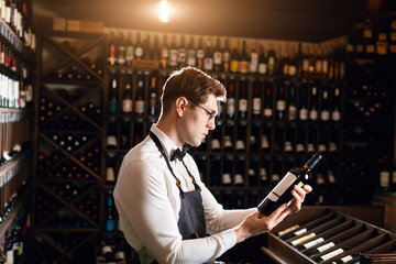 Professional male cavist examines the bottles with wine in wine shop, holding wonderful sample, ready to speak out about this wine to customers - obrazy, fototapety, plakaty