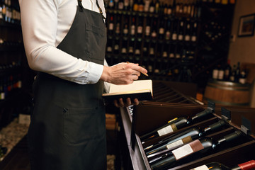 Professional male cavist examines the bottles with wine and making notes at his notepad about their features and origin in winery shop where people can buy a good wine.