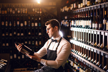 Handsome and young elegant brown haired cavist or wine seller with a bottle of wine on the background of dark wine house with shelves of bottles with alcohol.