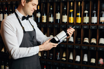 Young caucasian cavist dressed in white shirt and bowtie working in big vine shop presenting a bottle of red wine to customer