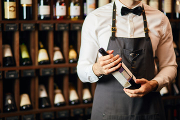 Cropped shot of wine seller or cavist in white shirt with bowtie and apron looking at bottle, telling about the origin of this sort of wine, wine shelves on the background - obrazy, fototapety, plakaty