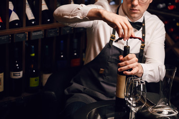 Young handsome sommelier pulling cork out of a bottle using a sommelier knife. Cropped photo of a waiter opening red wine bottle with corscrew in restaurant - Powered by Adobe