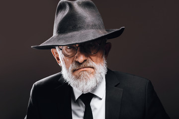 Old-aged bearded man in image of English secret agent wearing black suit with hat on his eyes posing against dark background.