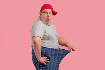 Happy surprised overweight man shows his big-size pants after keeping diet for three weeks isolated over pink background. Dieting concept.