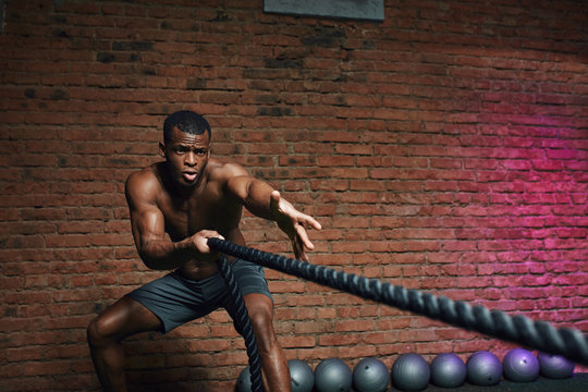 African man with naked torso doing fitness workout in cross fit gym. Shirtless strong athlete being concentrated on the exercise with battle rope requiring increased effort and strength.
