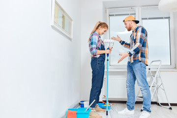 Fototapeta premium Newlyweds quarrelling because of renovation in new apartment. Pissed off irritated young man scolding his embarrassed guilty wife standing in white room with set of paint tools