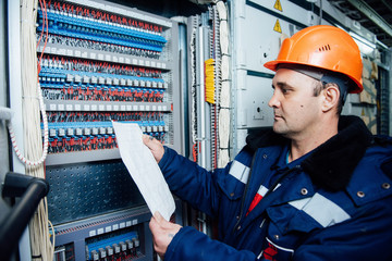 A man in overalls wearing a protective helmet is standing by an electrical shield.