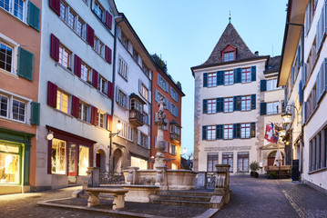 Z&uuml;rich Napfbrunnen beim Napflatz an der Spiegelgasse, Abendd&auml;mmerung, Z&uuml;rcher Altstadt, Brunnenturm, mittelalterlicher Wohnturm, Schaufenster, Beleuchtungen, blauer Abendhimmel, Schweizer Fahne