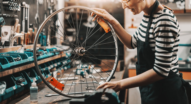 Cute Caucasian female worker holding and repairing bicycle wheel while standing in bicycle workshop.