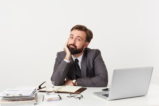 Indifferent office worker with beard sits at the table props his chin with his hand looks camera, feels bored apathetic uninterested, dressed in a suit with a tie, over white background