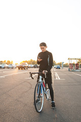 Portrait of a young rider standing with a bike on the background of the sunset and using a smartphone. Cyclist is cycling and using the internet on the smartphone.