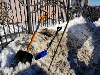 Snow shovel and ice ax in the snow against the background of a tile and fence in spring or winter