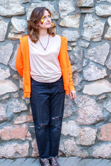 A young woman in full growth against a stone wall. A girl in a smart orange dress next to a wall of...