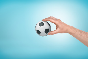 Male hand holding small football ball between fingers on blue background